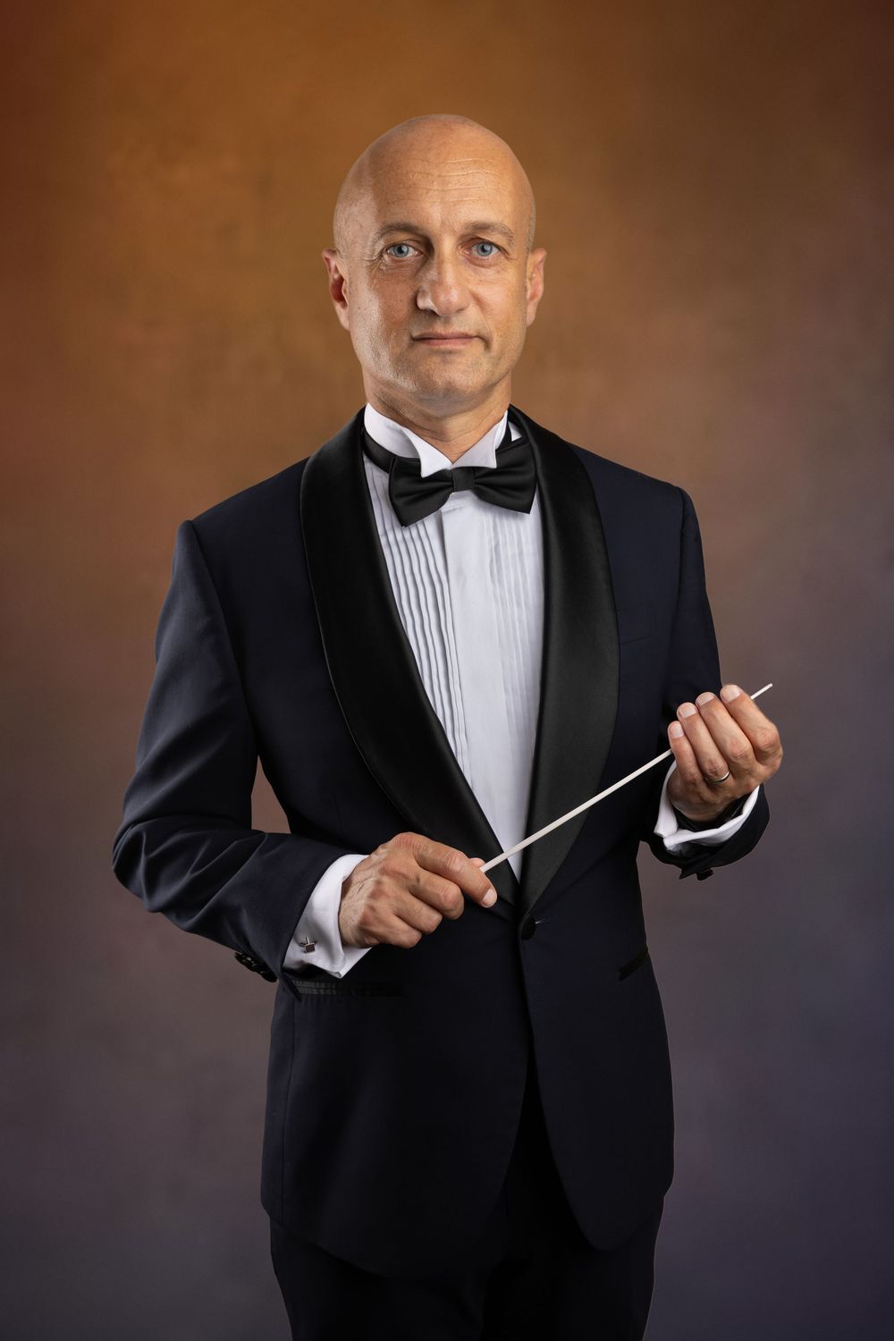 Bald male orchestra conductor in black tuxedo and bow tie holding a conducting baton against brown background.