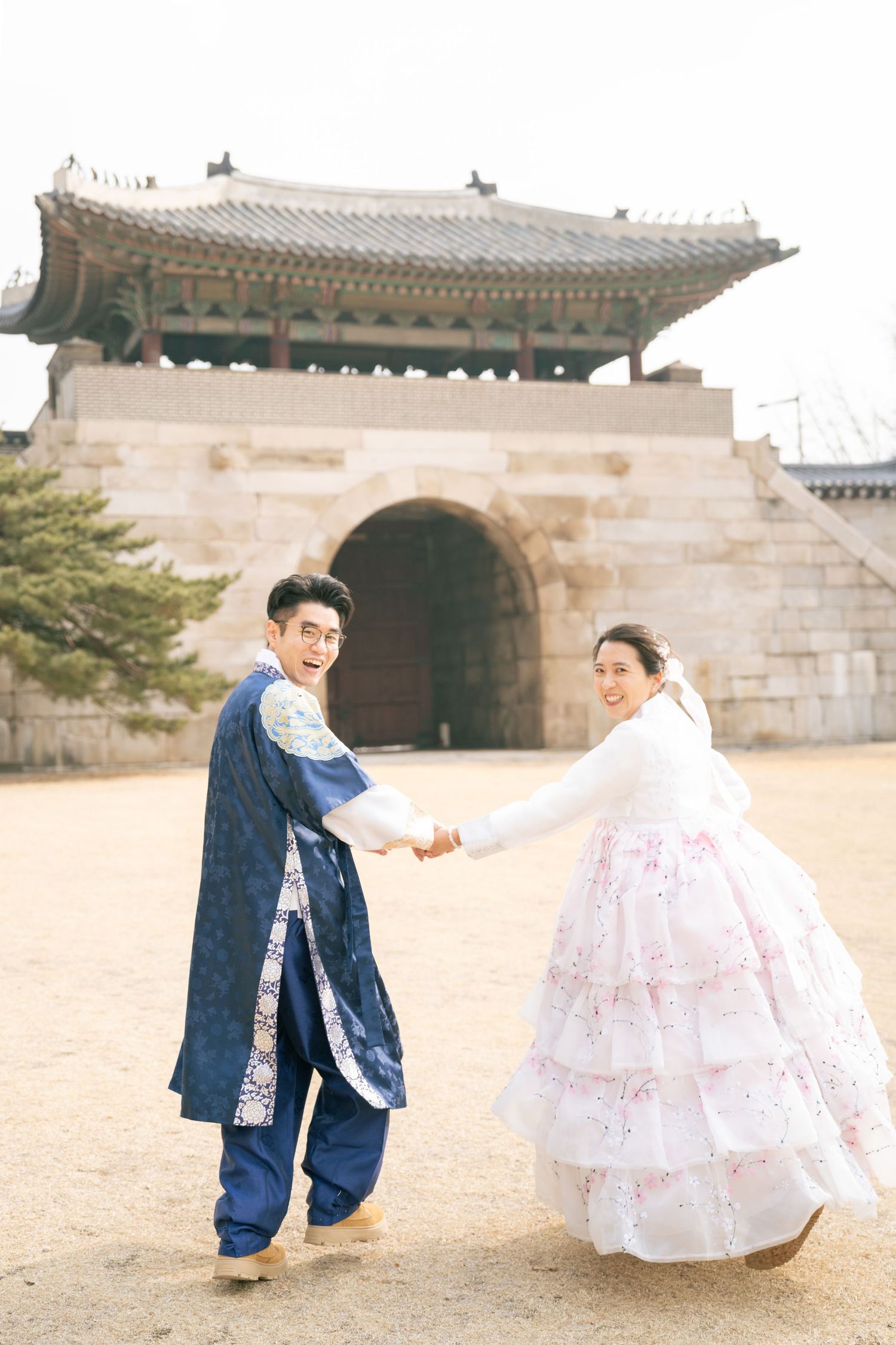 A couple in traditional Korean wedding attire poses playfully in front of historic palace architecture.
