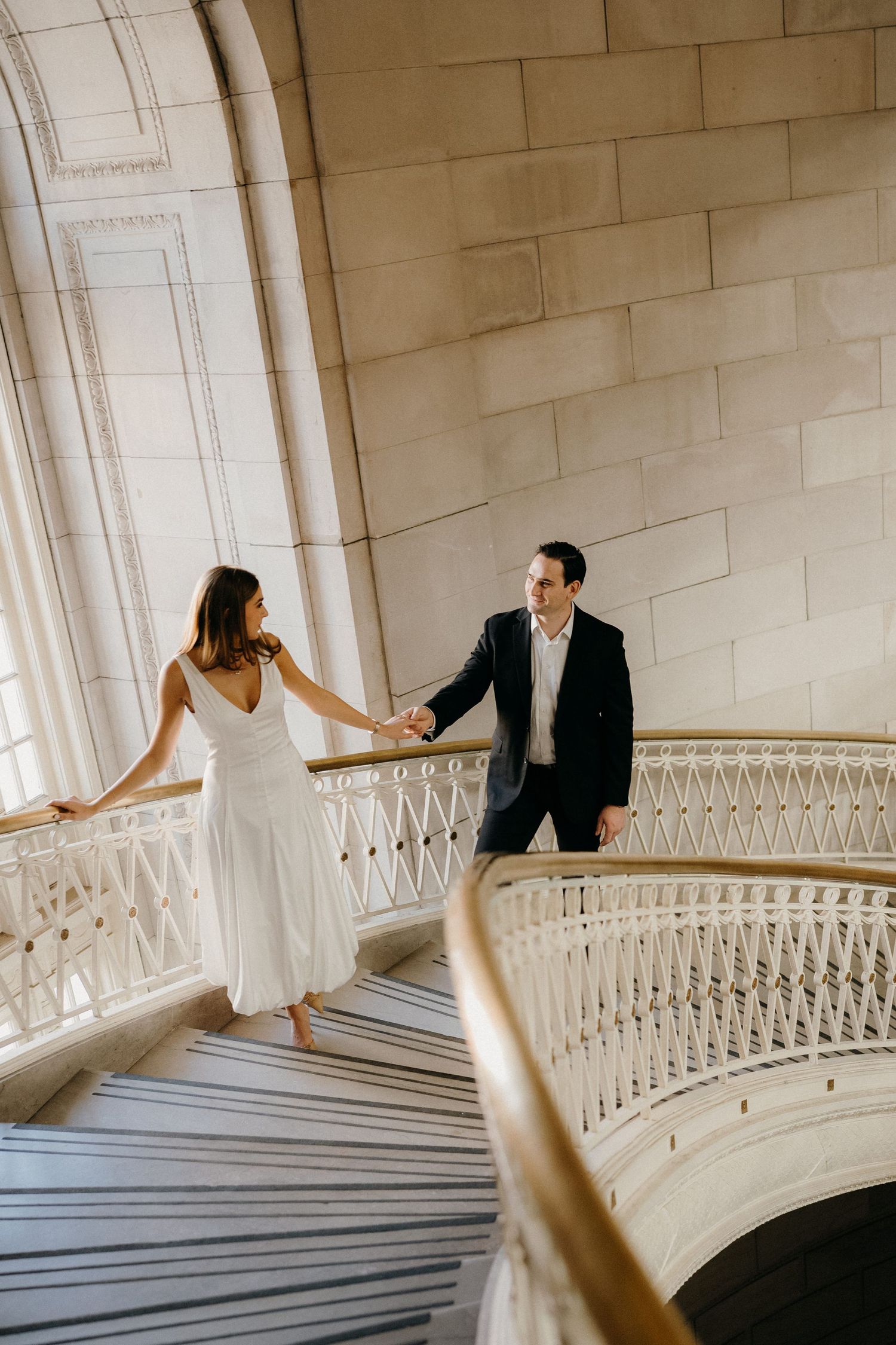 Elegant couple stands on a grand marble staircase with ornate railings in a classic architectural setting.
