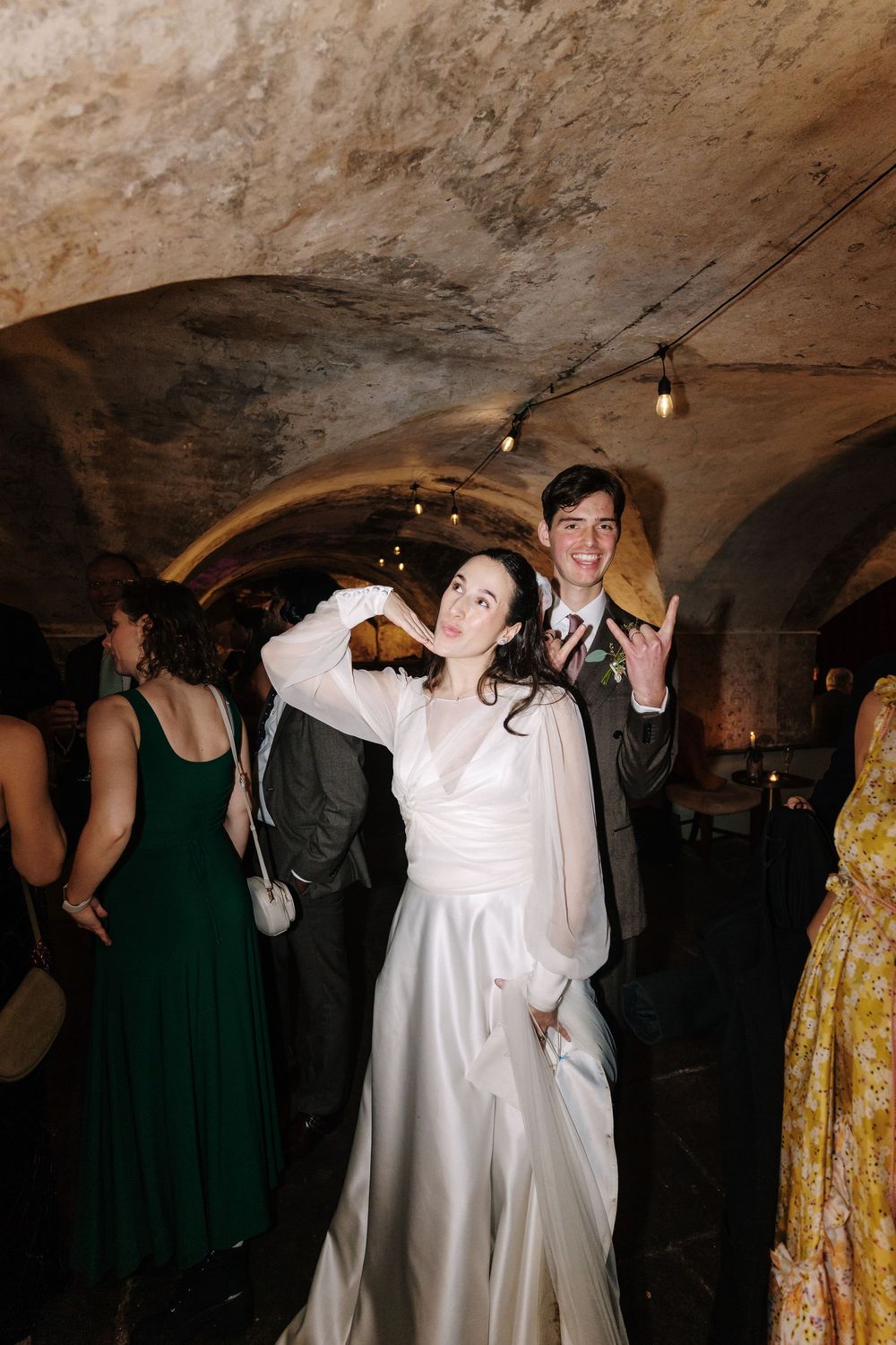 Wedding guests dancing and celebrating in a dimly lit cave or underground venue with stone walls and arched ceilings.