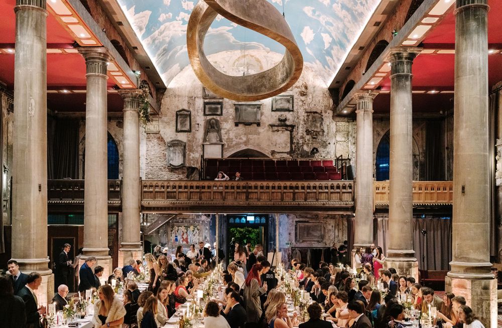 A panoramic view of an ornate historic dining hall with vaulted ceilings, columns and guests seated at long tables.