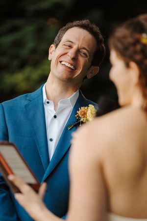 A groom in a blue suit jacket and white shirt smiles warmly during an outdoor wedding ceremony.