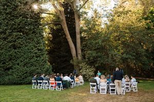 Outdoor wedding ceremony setup with white chairs arranged under tall trees in a garden setting.