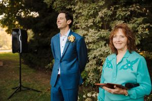 A couple in coordinating blue suit and turquoise blazer stand together outdoors in a garden setting.