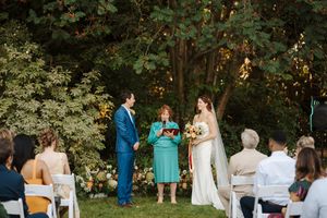 Outdoor wedding ceremony in a wooded area with guests gathered on white chairs watching the proceedings.