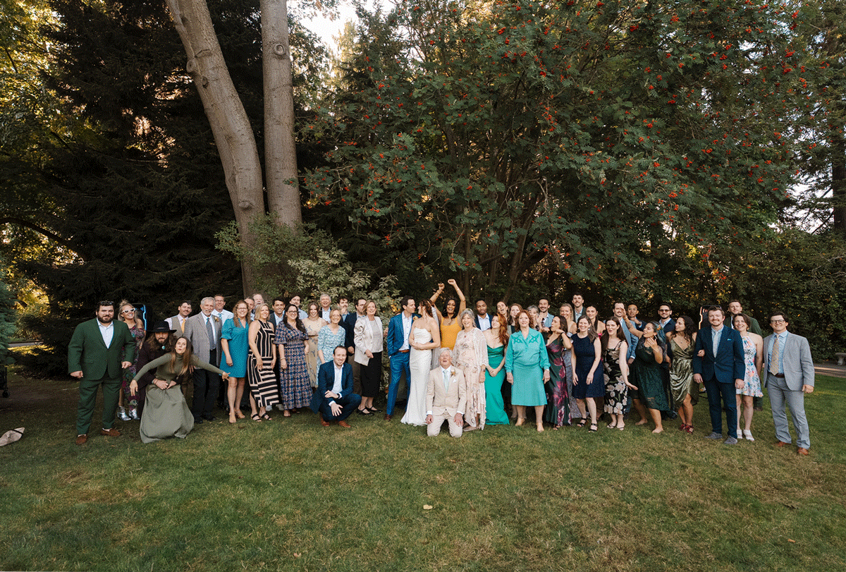 Large group photo of wedding guests gathered outdoors on a lawn with trees in the background during golden hour.