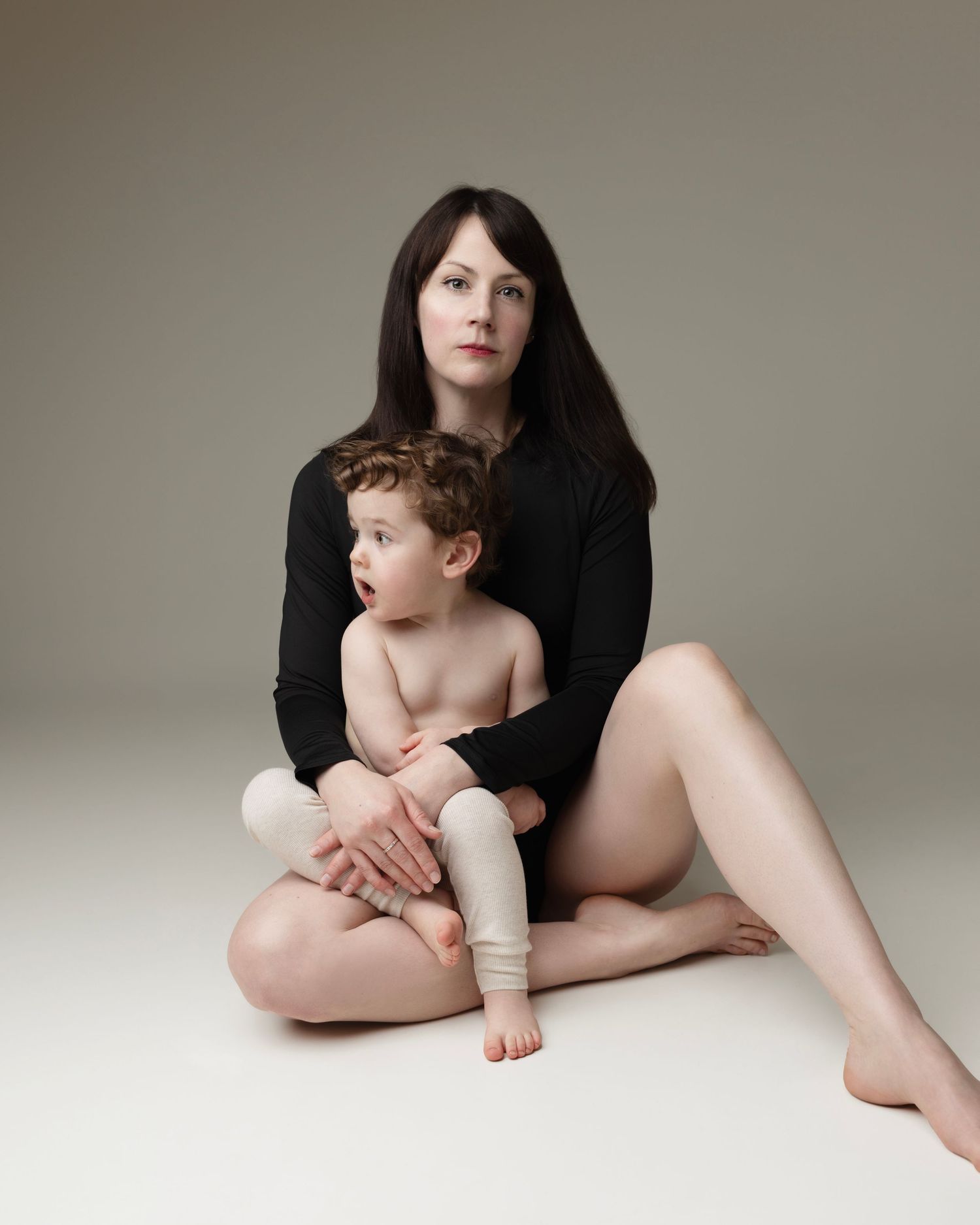 Artistic studio portrait of parent and toddler in minimalist setting with black sweater against neutral background.
