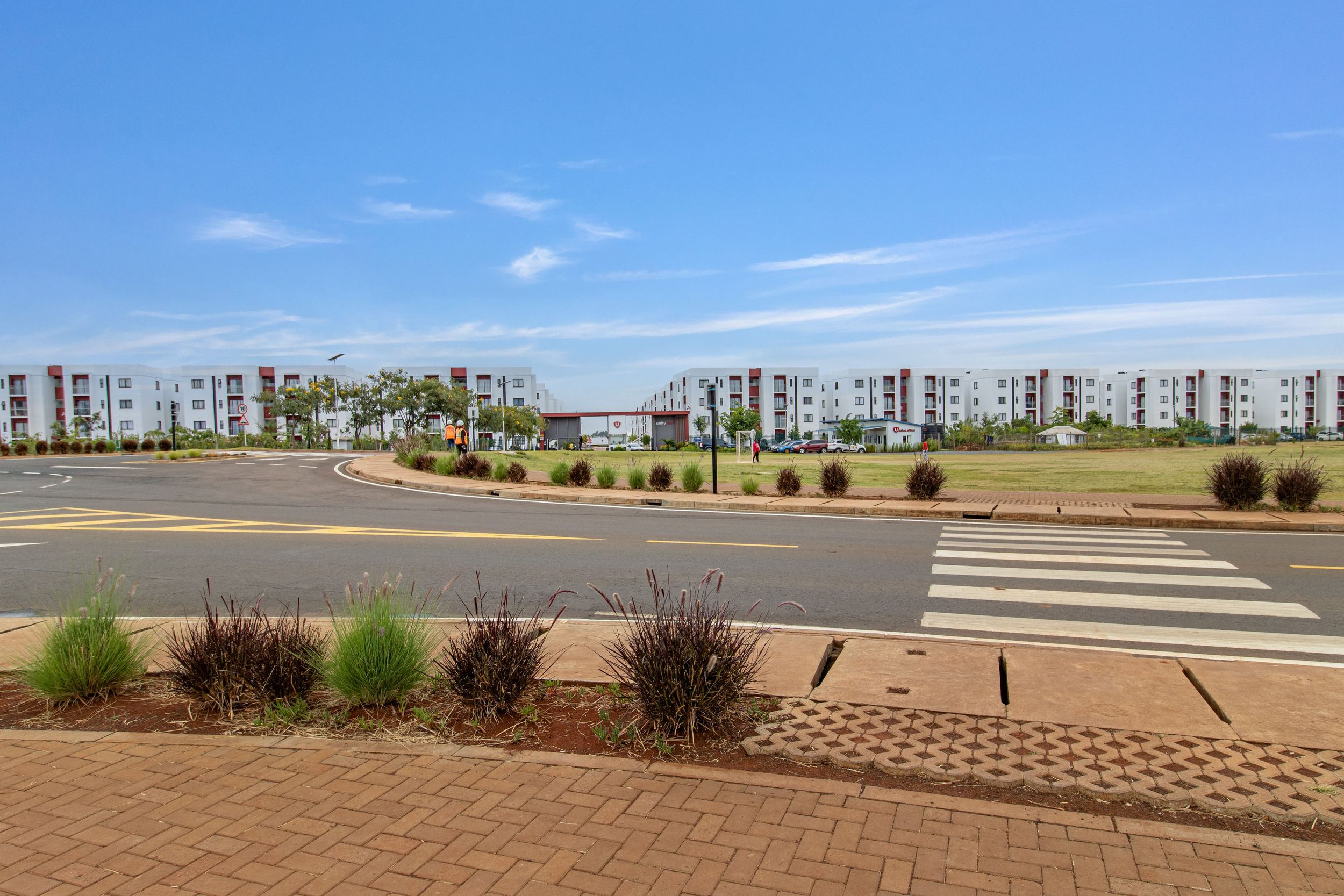A row of modern apartment buildings and palm trees line a street under a bright blue sky with scattered clouds.