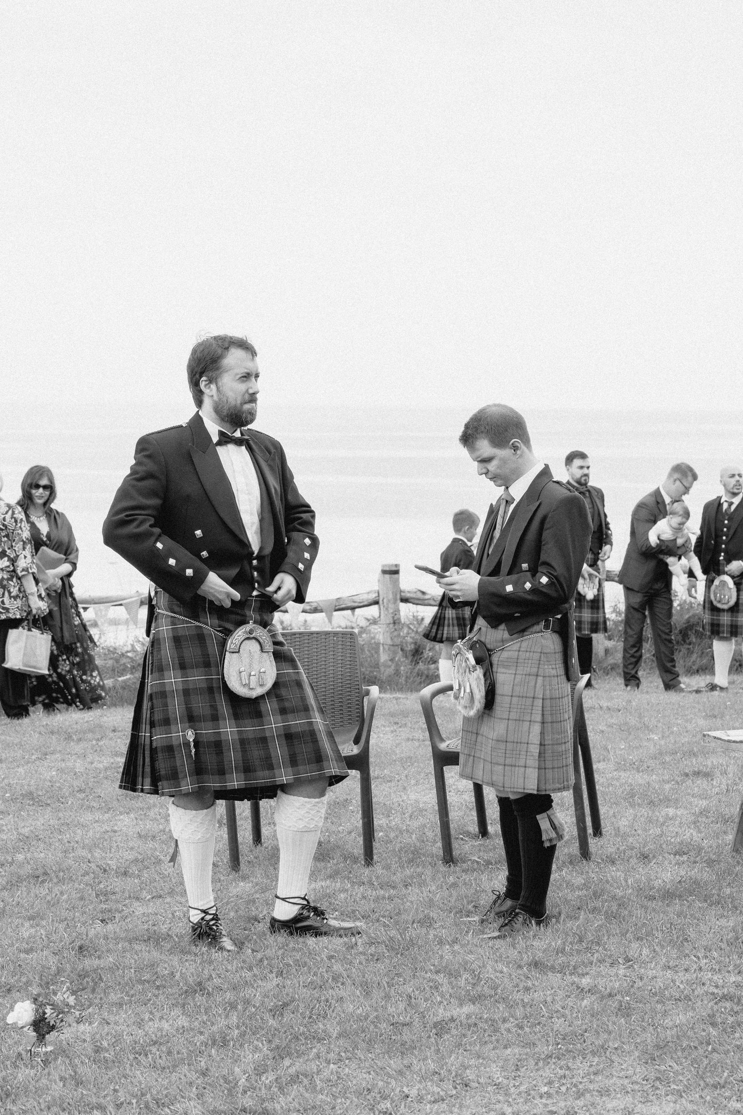 Men in traditional Scottish kilts gather on a grassy field during an outdoor event in black and white photography.