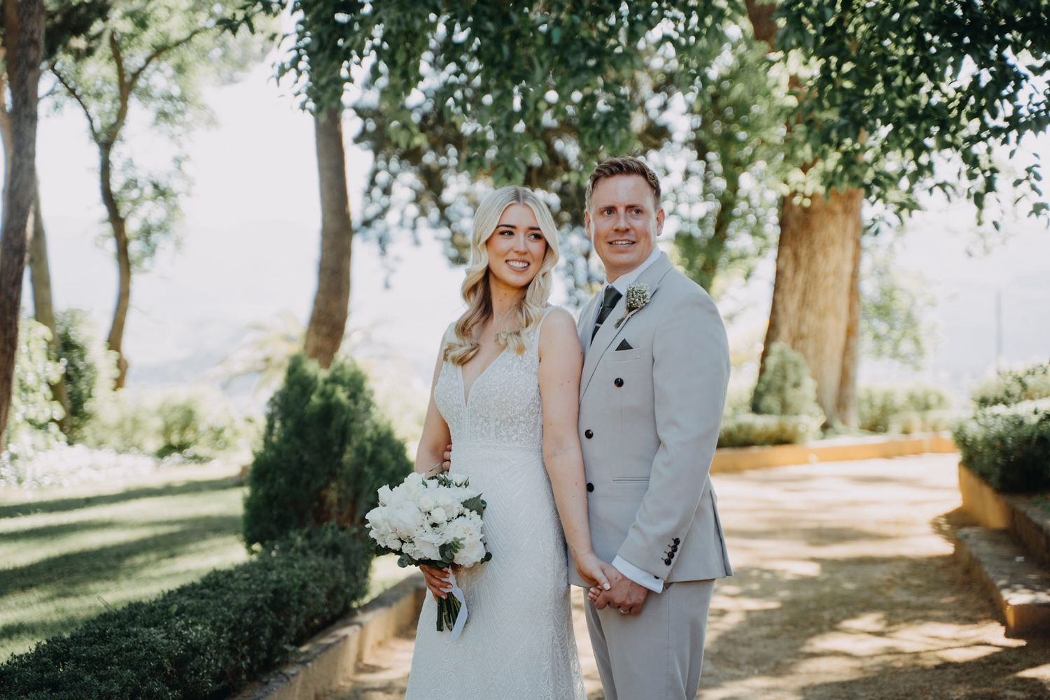 A couple in wedding attire walks together through a sunlit garden path with trees creating a natural archway.