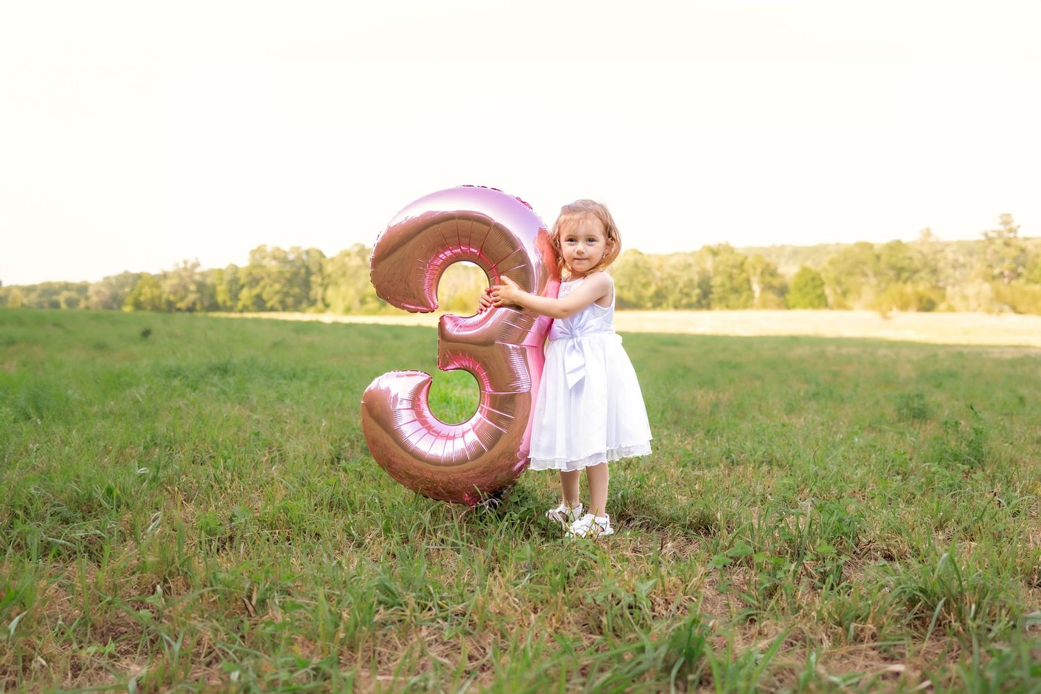 A pink number 3 balloon stands in a sunny grassy field with someone in a white dress celebrating a birthday.