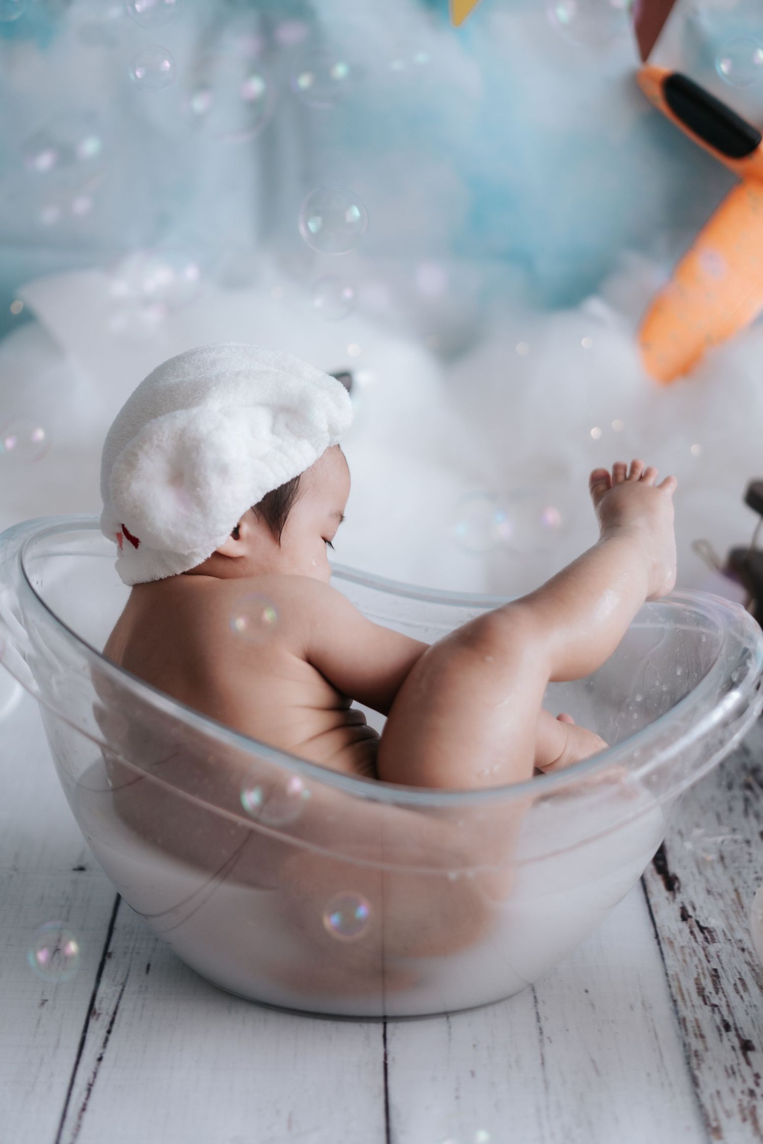 A baby doll taking a bath in a white ceramic tub with soap bubbles floating in the air.