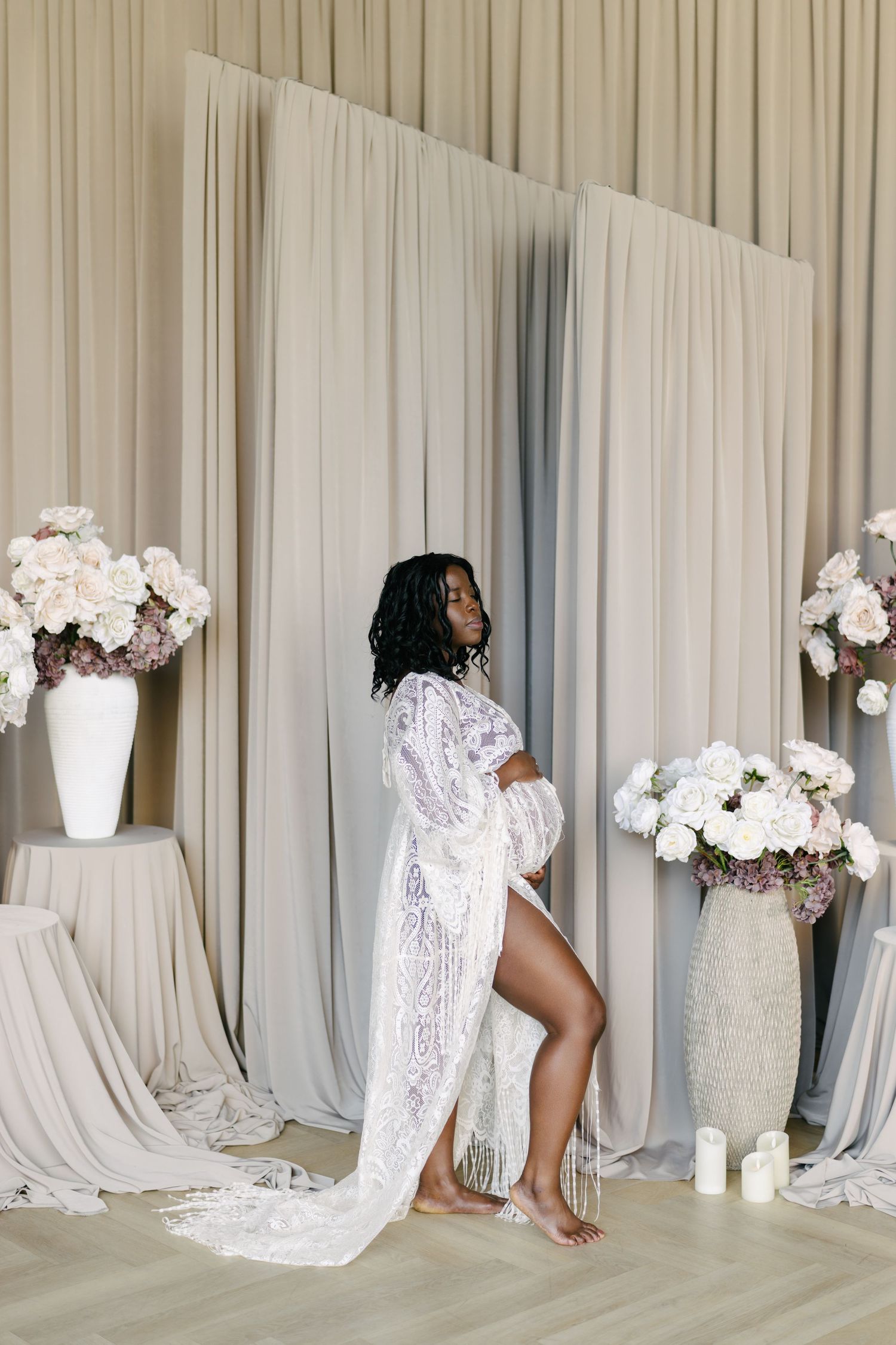 Expectant mother in a lace robe posing in a studio between white floral arrangements.