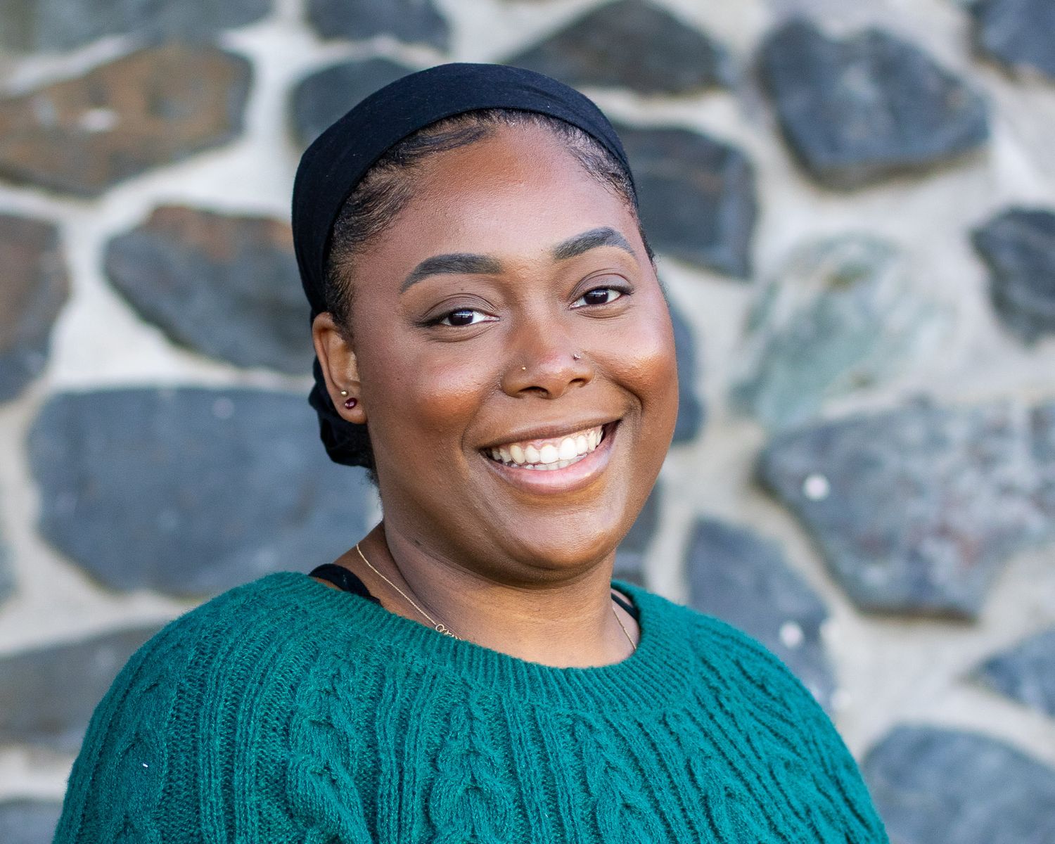 A person in a teal sweater smiles warmly against a stone wall background.
