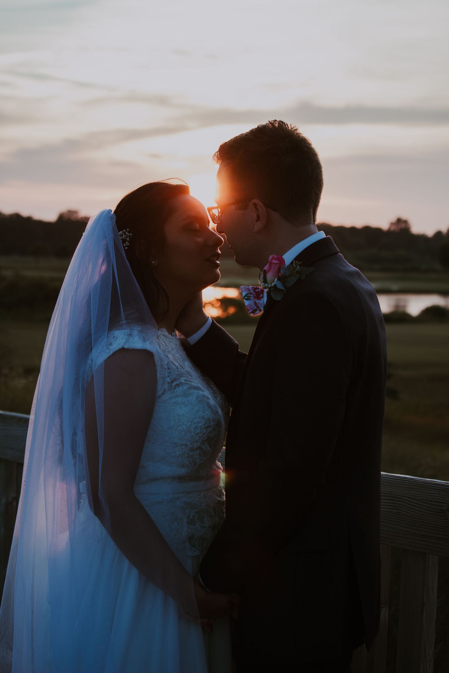 Romantic silhouette of bride and groom almost kissing at sunset with scenic waterfront background.