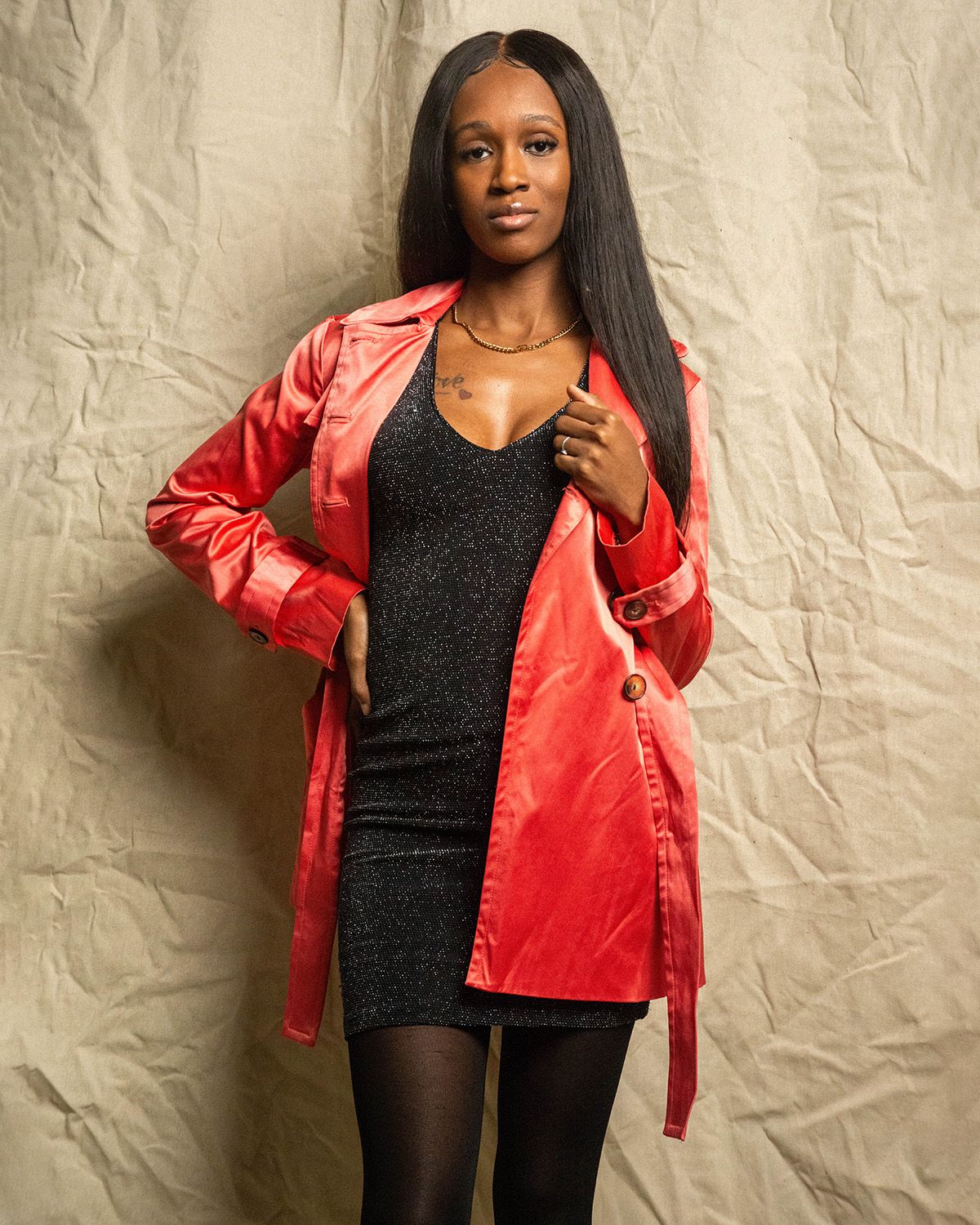 Fashion model poses in a black top and red satin jacket against a textured beige wall.