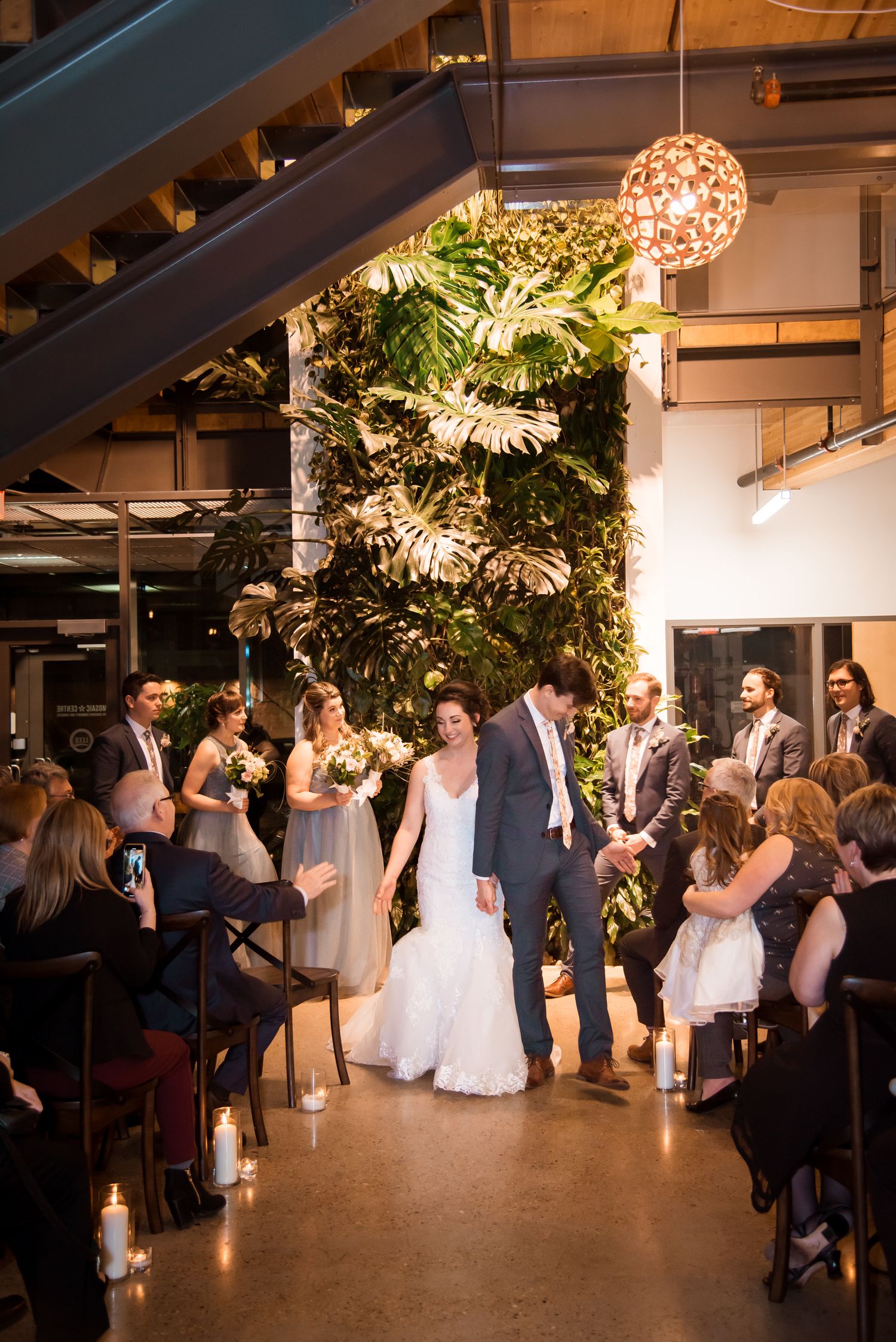Wedding reception guests dance and celebrate in an industrial-style venue with hanging floral lights and greenery decorations.