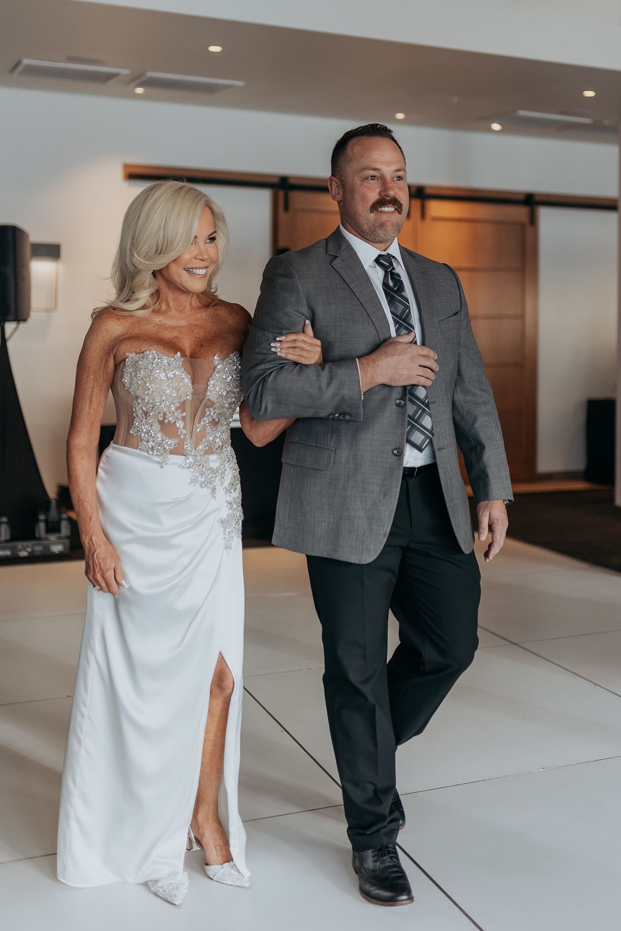 A bride in a white gown and her brother in a suit walk down the wedding aisle together on a polished floor at an indoor venue.