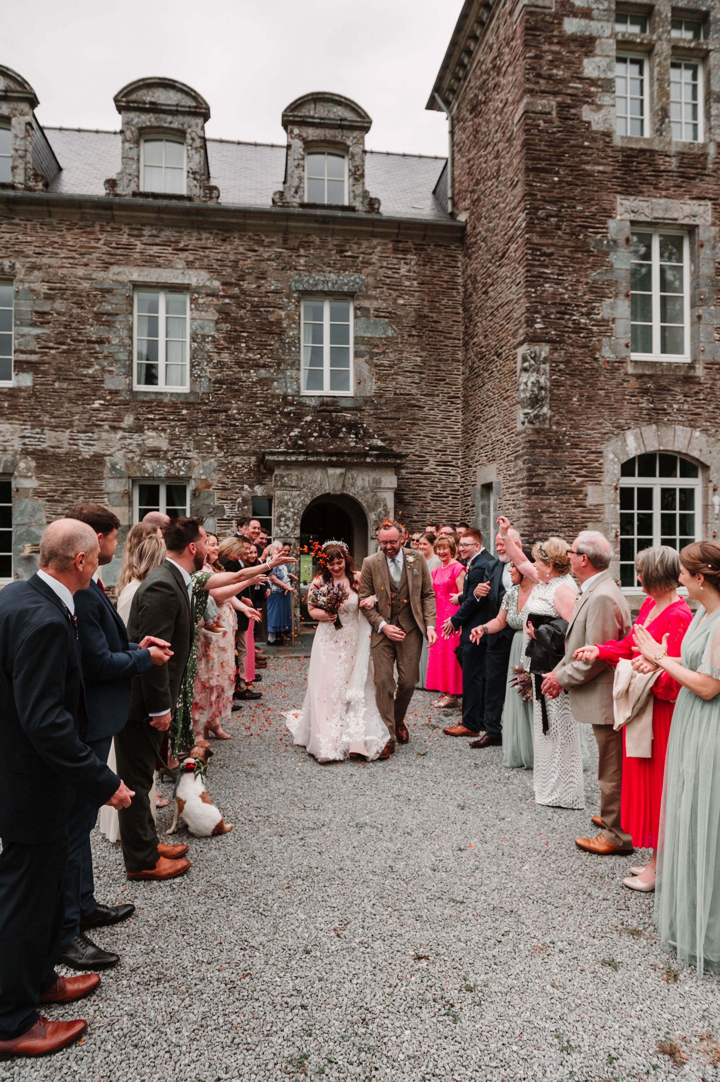 Sortie des mariés devant un château en pierre en Bretagne, invités lançant des confettis lors de la cérémonie.