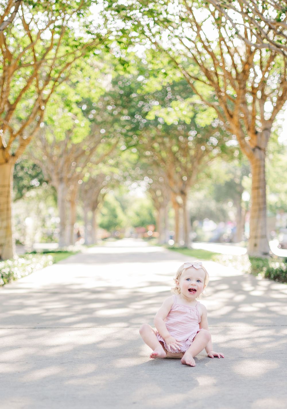 A toddler in pink sits laughing on a tree-lined path on a sunny day.