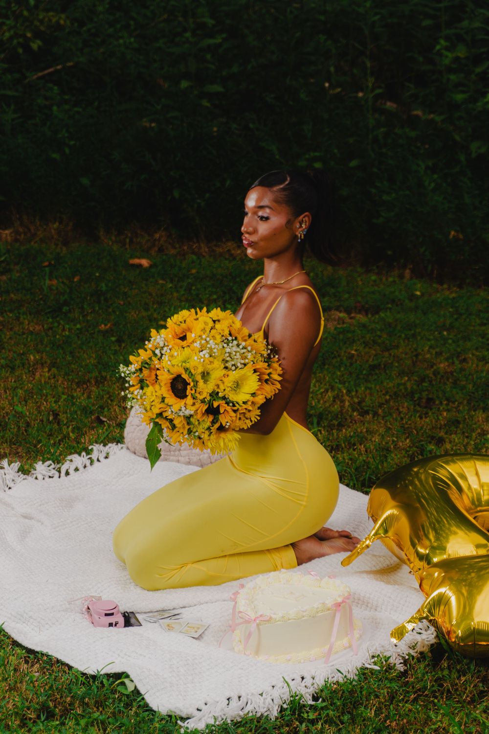 A figure in a yellow bodysuit reclines on white fabric outdoors with sunflowers in a romantic garden photoshoot.