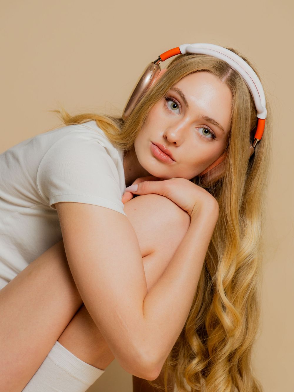 Portrait of someone wearing a white t-shirt and white and orange headphones with long blonde wavy hair against beige background.