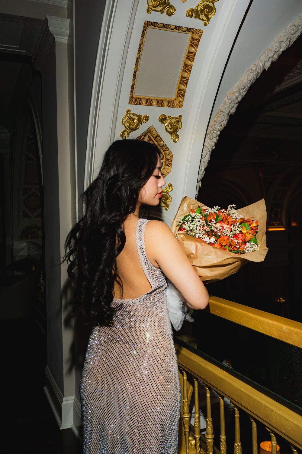 A person in a sparkly silver dress poses dramatically against an ornate wall with gold accents.