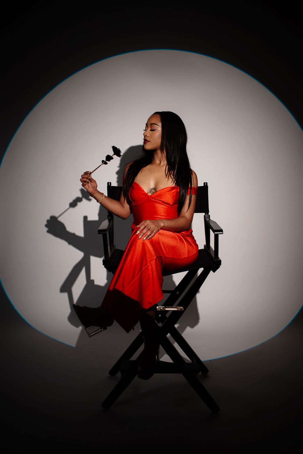 A person in a red dress sits on a director's chair in dramatic spotlight lighting against a gray background.