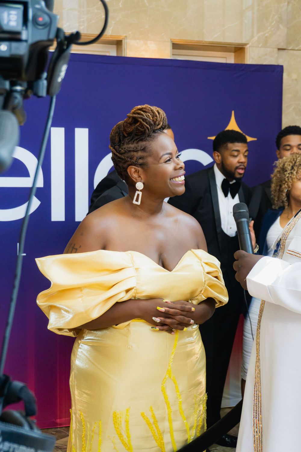 A person wearing a stunning yellow off-shoulder gown smiles at an event with a blue backdrop.