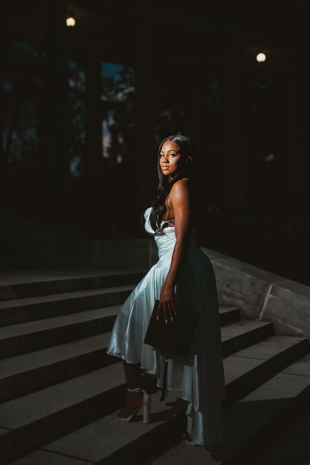A person in an elegant light blue dress poses on concrete steps at dusk with dramatic lighting and shadows.