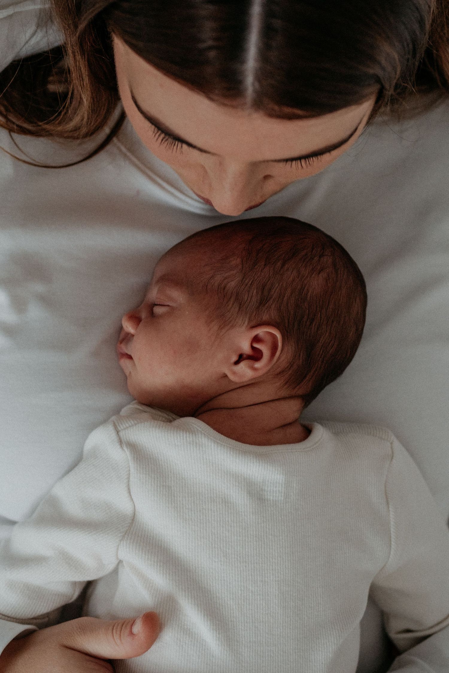 Tender newborn portrait of a mother kissing her baby while lying together, capturing a peaceful and emotional early bonding moment.