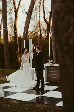 Joyful bride and groom dance together on checkered outdoor dance floor at golden hour autumn wedding.
