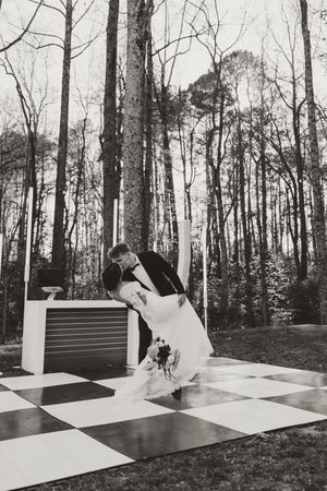 Black and white photo of groom dipping bride in romantic kiss on checkered dance floor in forest setting.