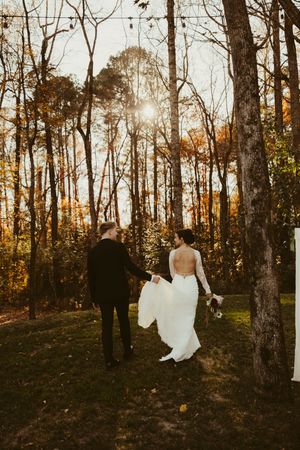 Bride and groom walk hand in hand through golden autumn forest at sunset after outdoor wedding ceremony.