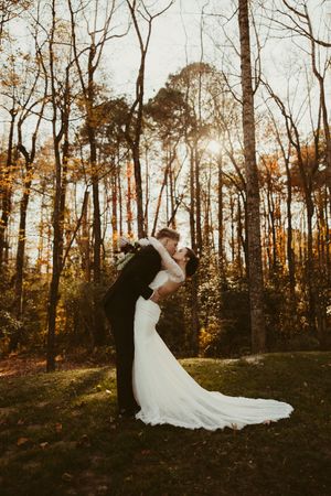 Groom dips bride for romantic kiss in sun-drenched autumn forest during outdoor wedding photo session.