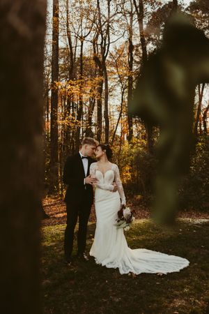 Elegant bride and groom embrace tenderly in golden autumn woodland, bride wearing lace long-sleeve gown.