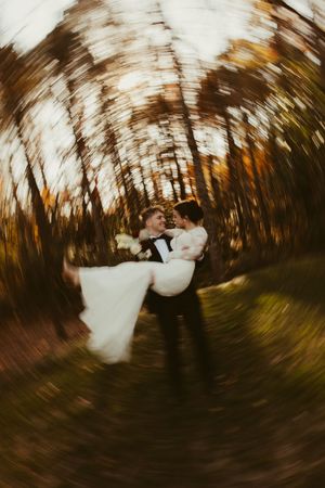 Groom lifts laughing bride in his arms in a dreamy zoom-blur photo among colorful autumn trees.