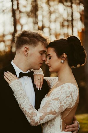 Bride and groom in elegant wedding attire share an intimate forehead touch outdoors in golden sunset light.