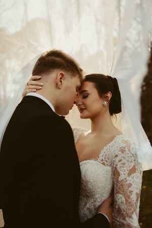Romantic bride and groom moment under flowing veil, touching foreheads in warm autumn light.