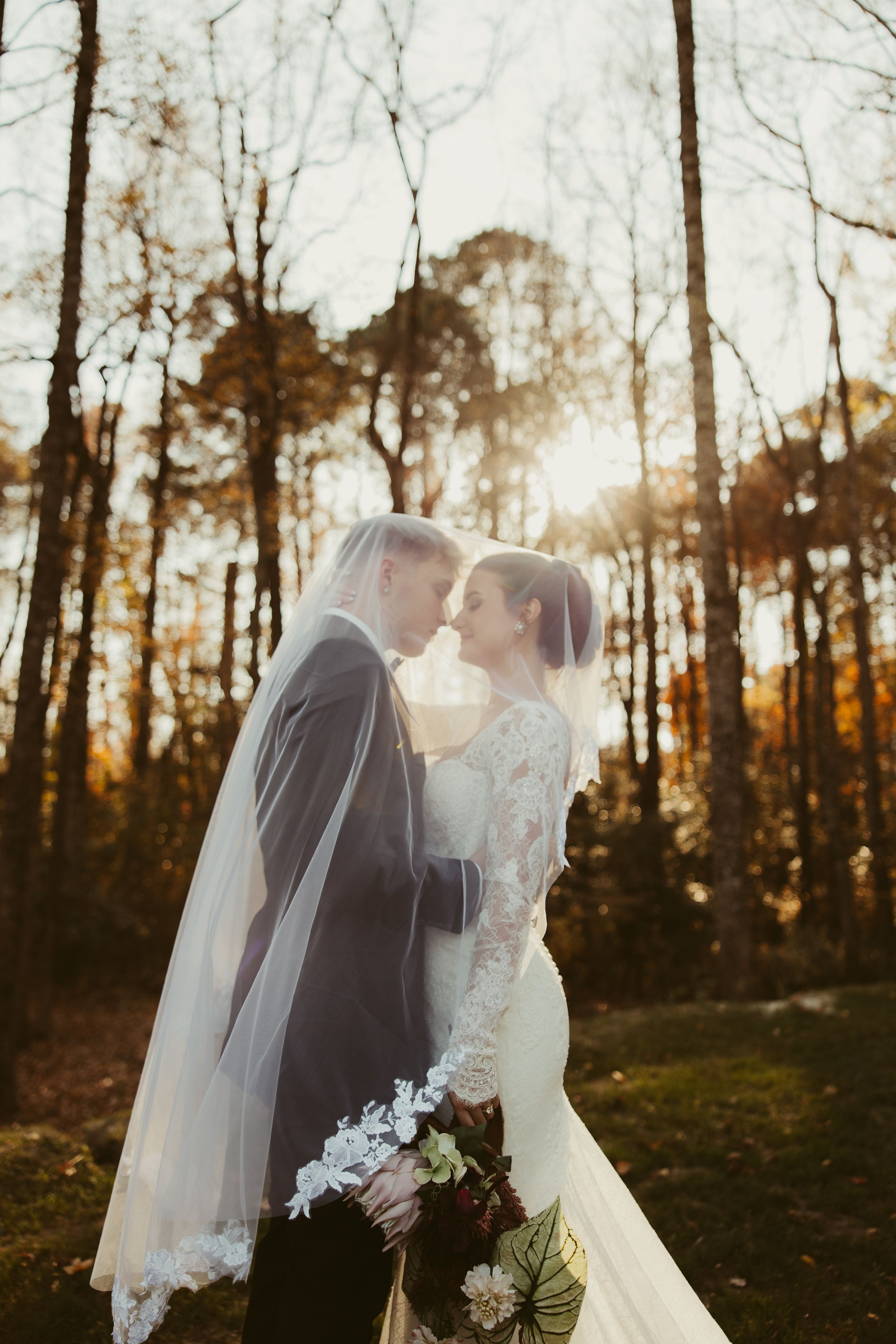 Bride and groom share a tender moment under a veil in a sunlit autumn forest setting.