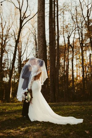Bride and groom kissing under a veil in a golden autumn forest, bride holding a bouquet.