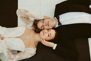 Bride and groom lying down together, bride in lace dress, groom in black suit, eyes closed.