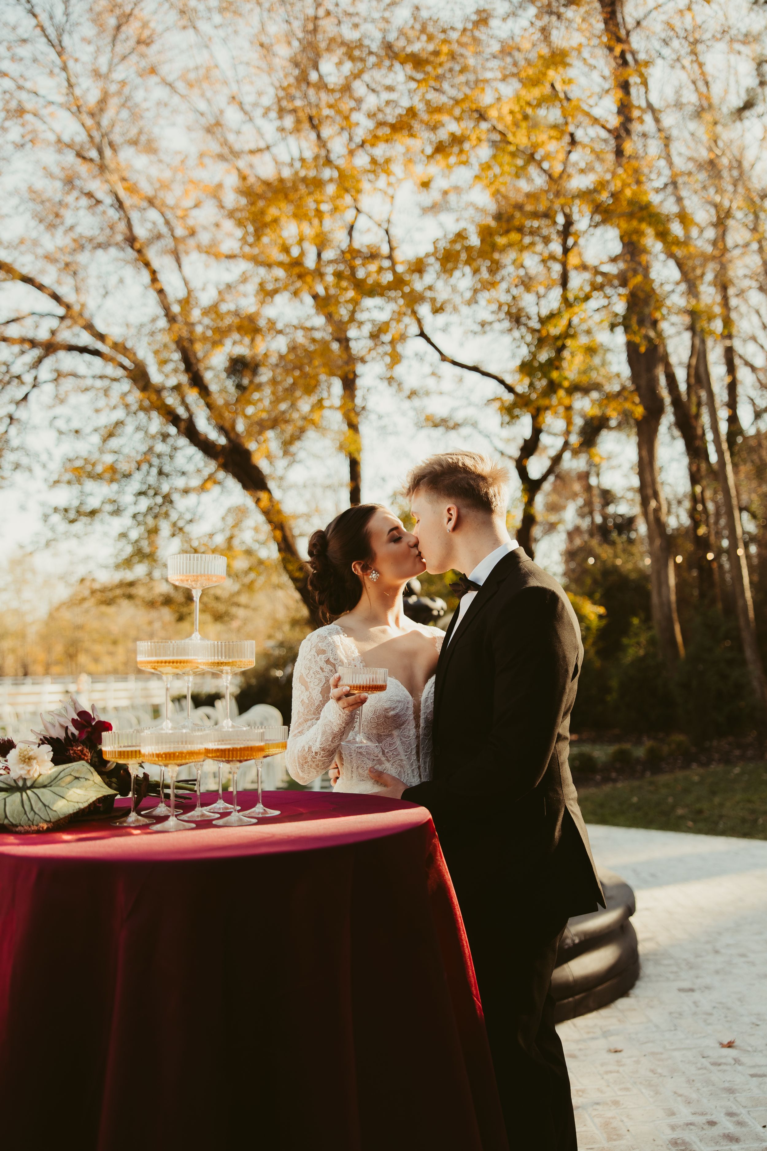 Bride and groom share a kiss beside champagne tower at outdoor autumn wedding reception table.