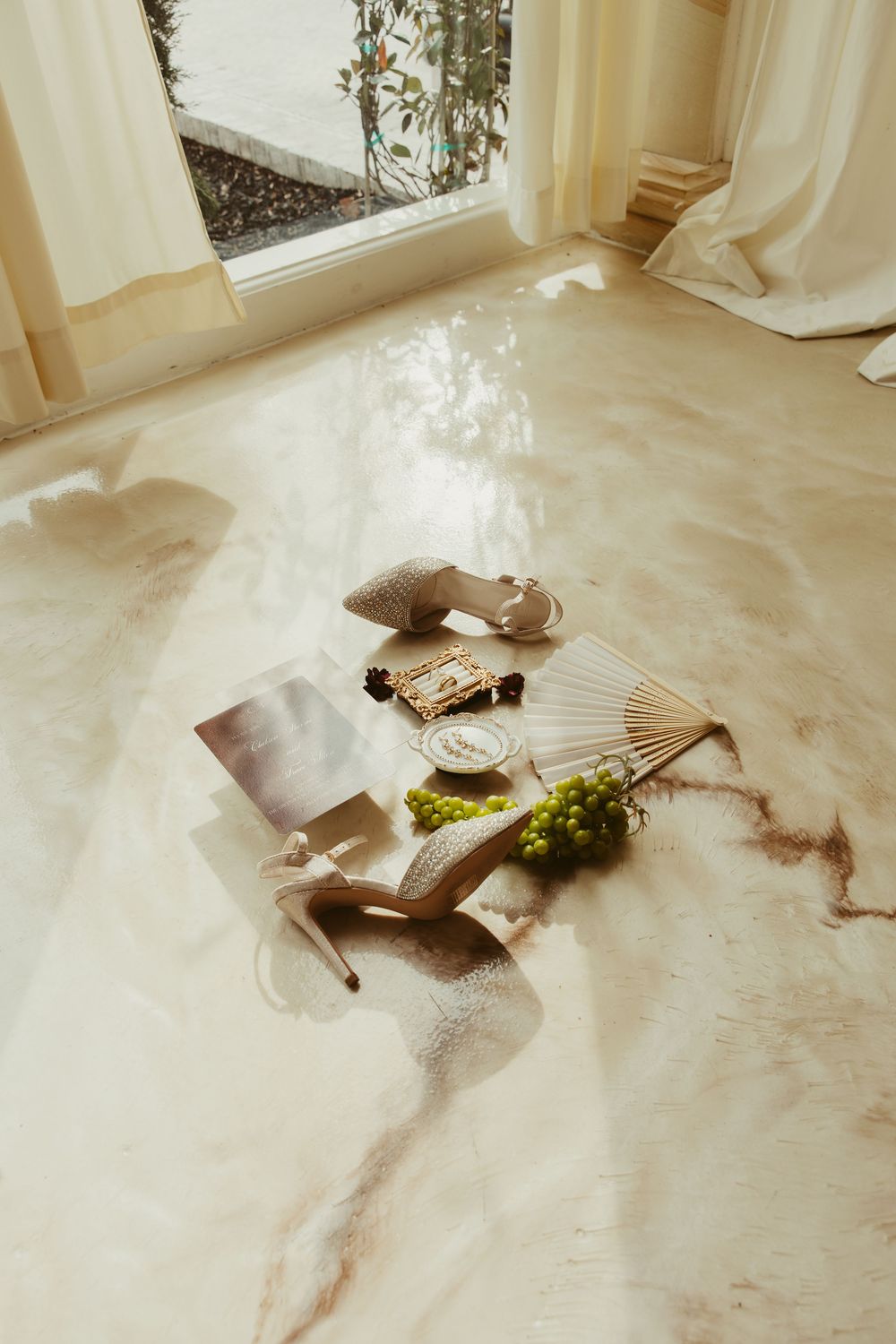 Elegant bridal flat lay with heels, fan, grapes, and accessories on marble floor near window.