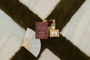 Wedding stationery flat lay featuring burgundy invitation, paper fan, and perfume on striped grass.