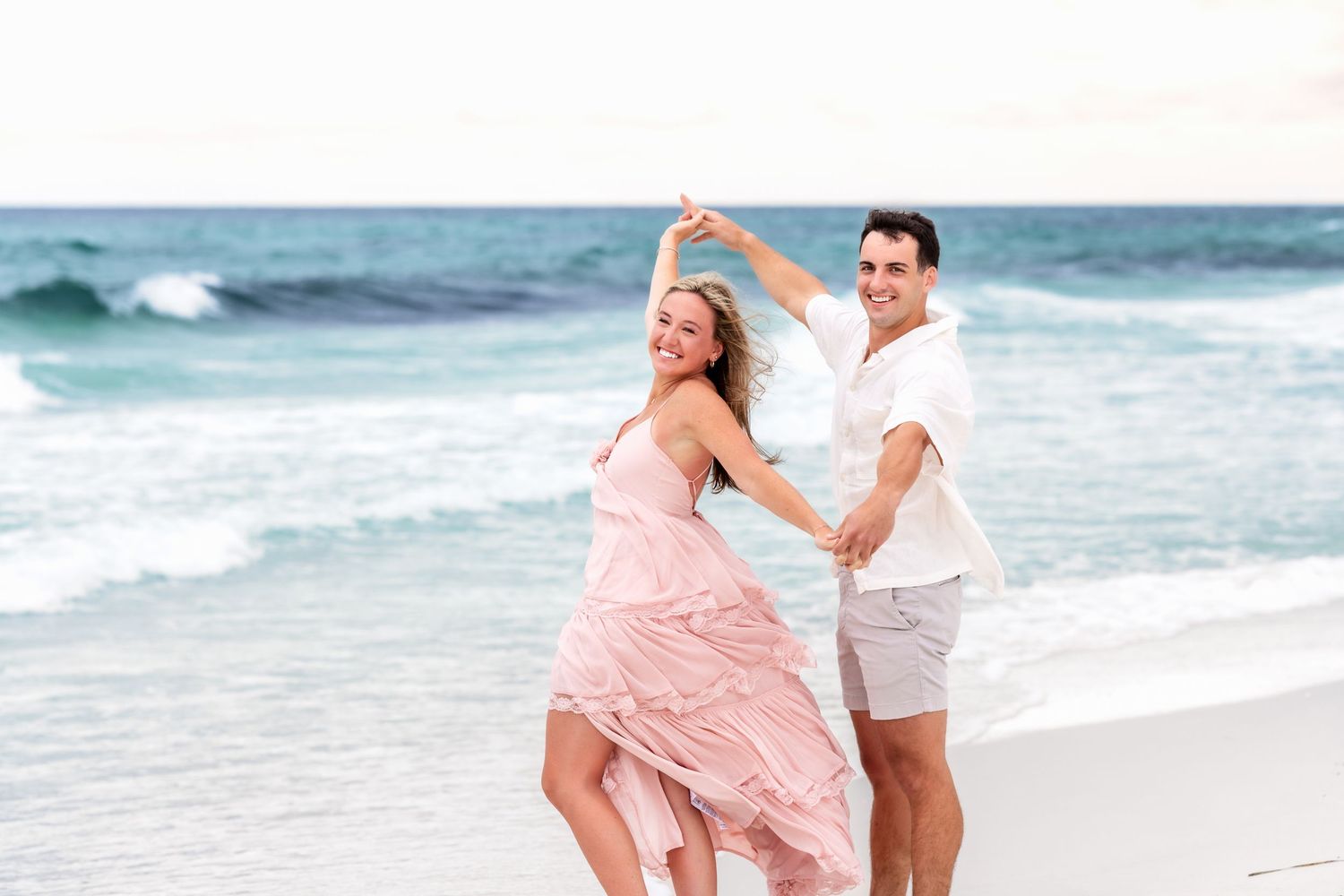 A couple in white and pink outfits dance playfully together on a sunny beach with ocean waves in the background.