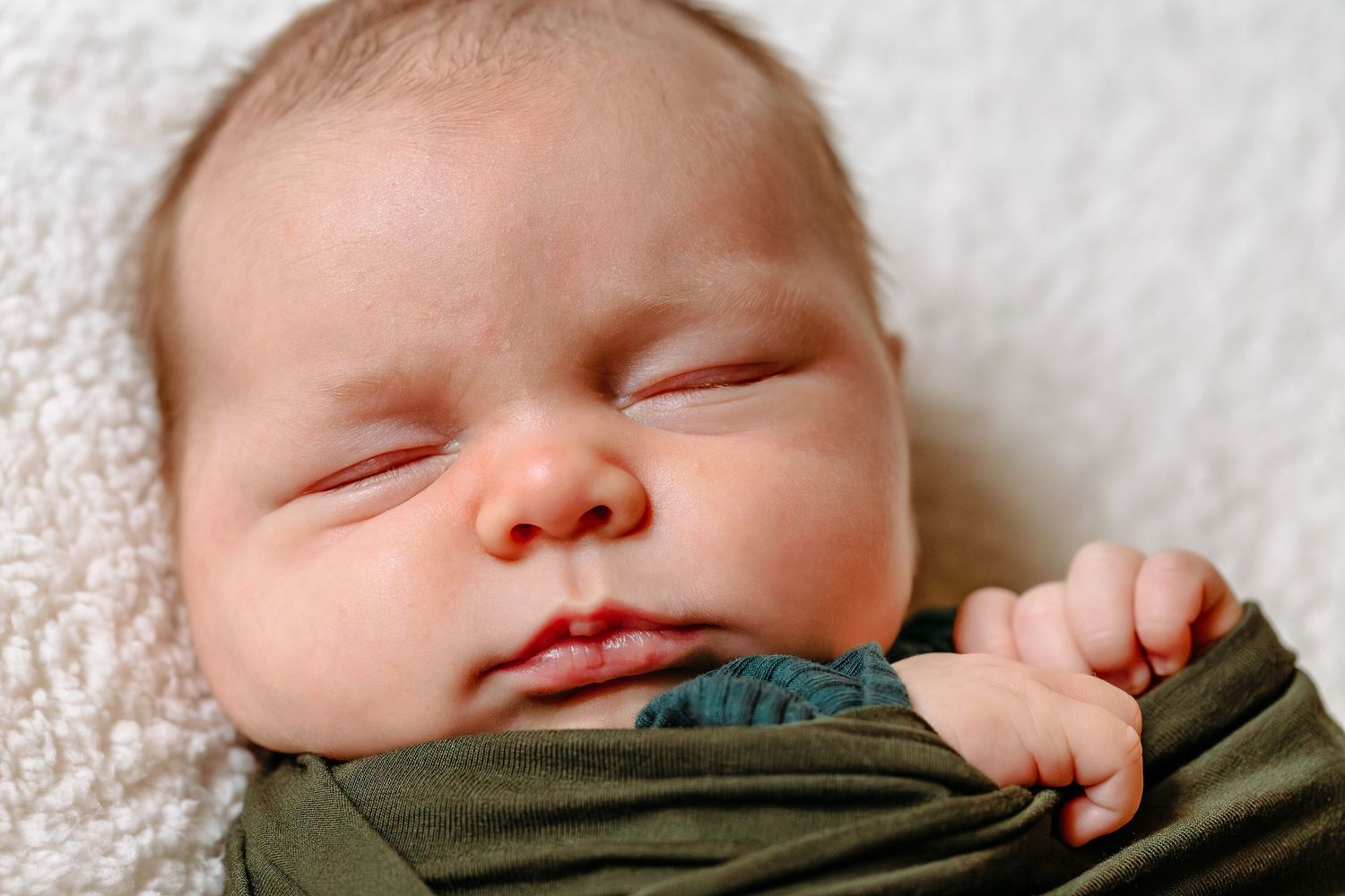 Sleeping baby wearing an olive green outfit rests peacefully on white textured bedding.