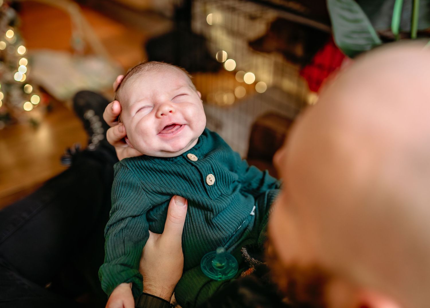 A laughing baby wearing a dark green button-up outfit with twinkling holiday lights in the background.