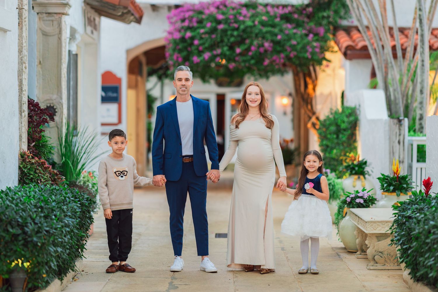 A family walks together on a garden path with white walls and purple bougainvillea flowers in the background.