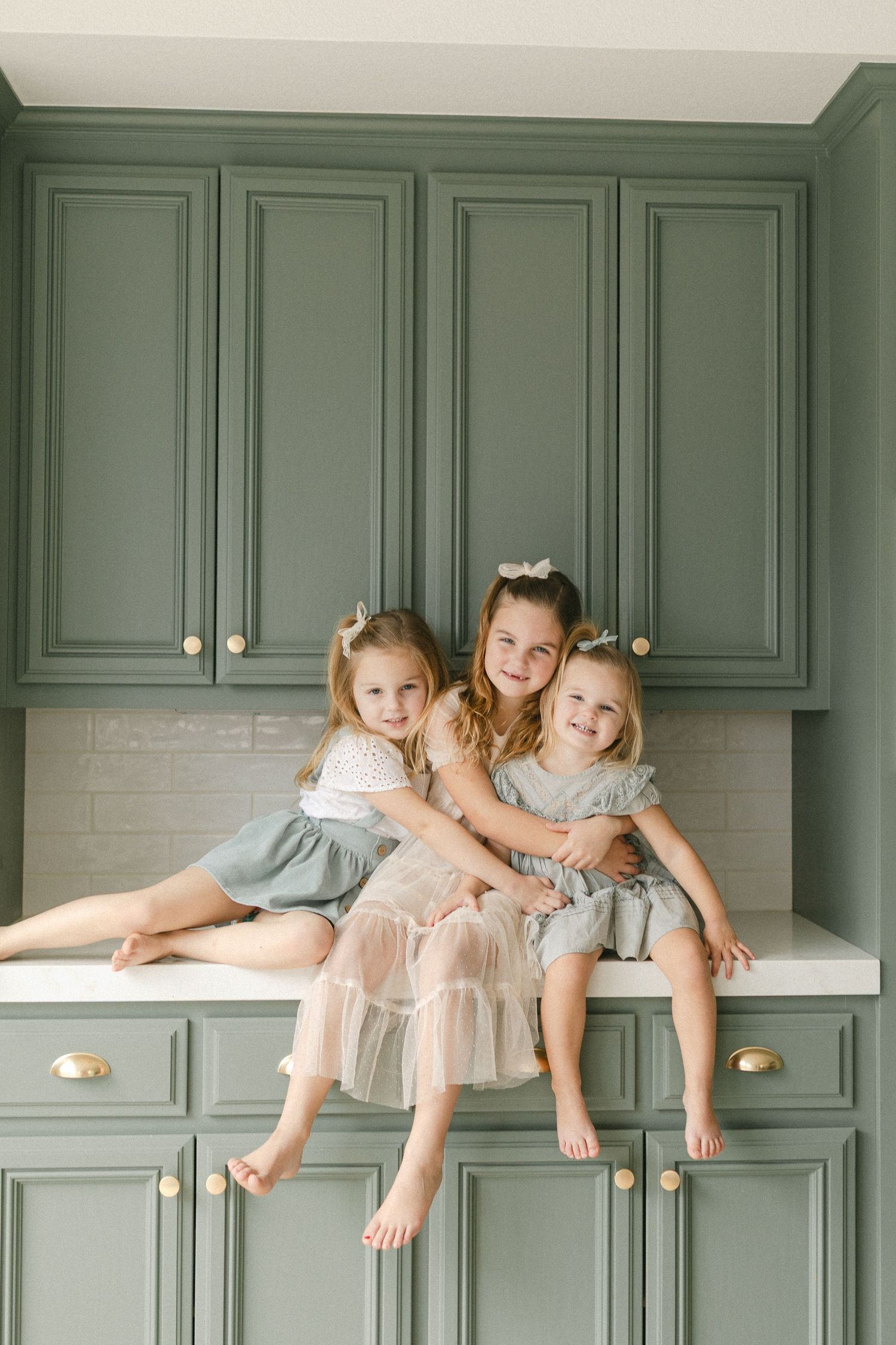 Child in gray dress playfully lounges on white kitchen counter with green cabinets and fresh fruit in background.